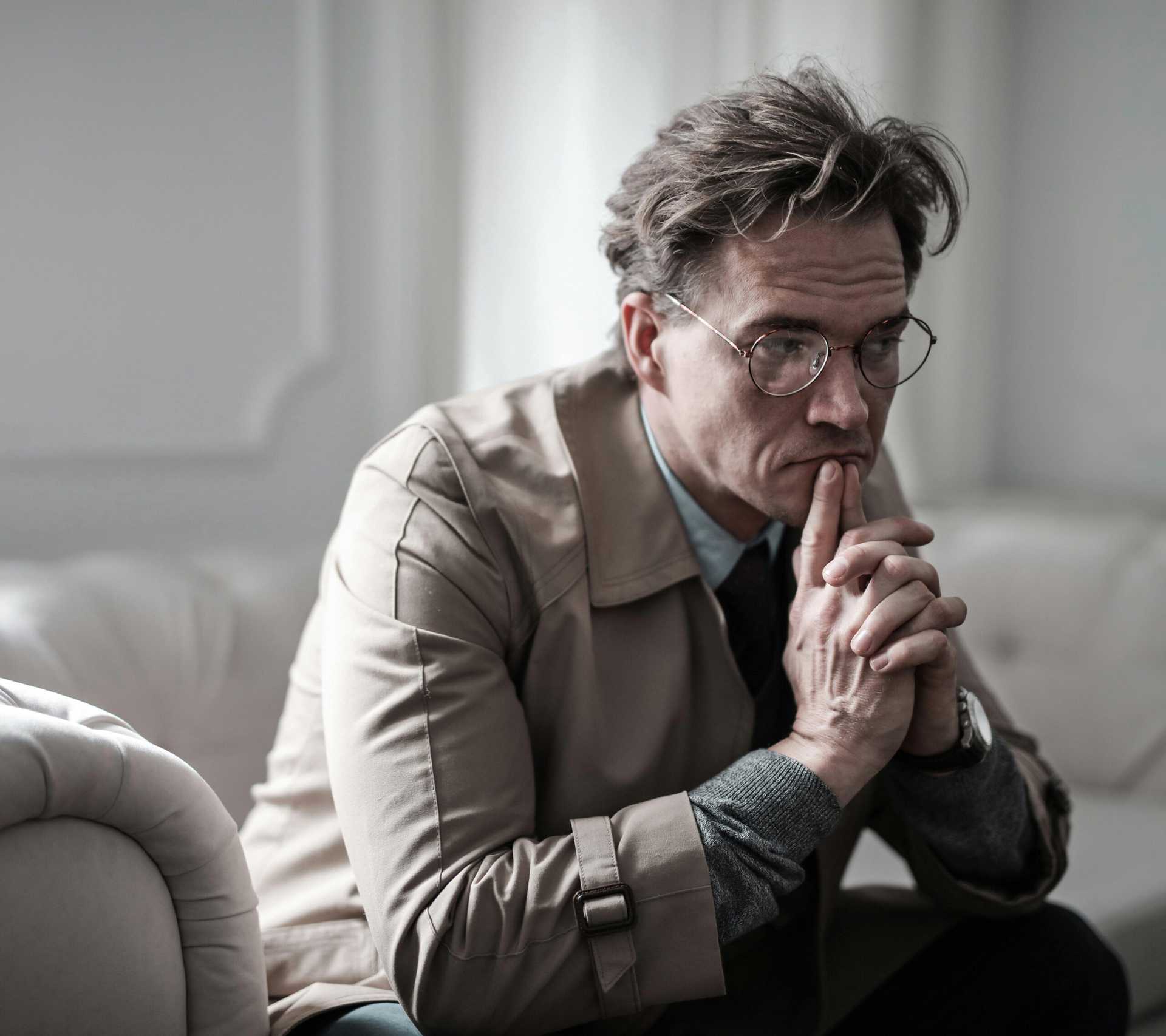 A man in a trench coat sits on a couch, leaning forward. He is deep in reflection.