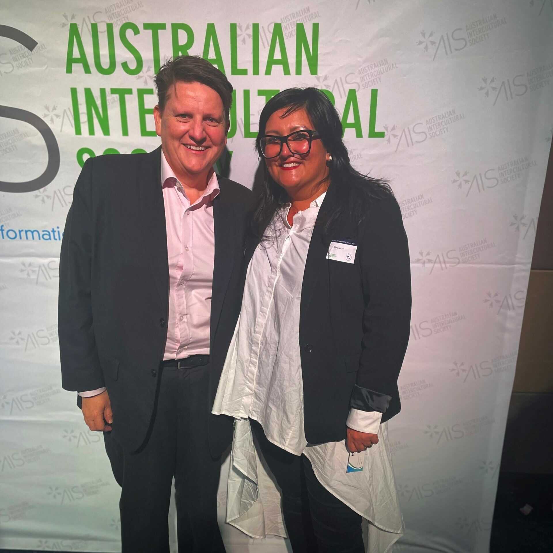 Ro Allen (left) and Taryn Lee (right) standing side-by-side, smiling in front of a banner which reads: Australian Intercultural Society
